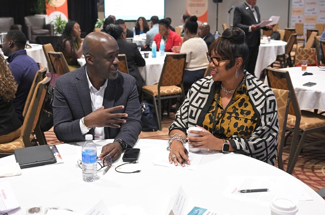 two people talking at a round table; other people occupy similar tables in the background
