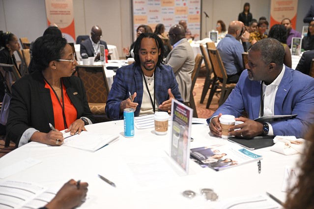 three people talk at a round table; others occupy similar tables in the background